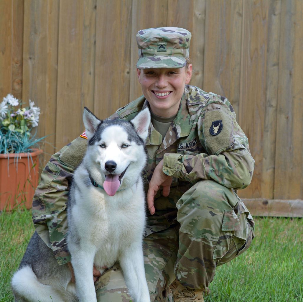 A woman in a military uniform kneeling next to a husky dog.
